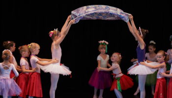 zwei kleine Mädchen tanzen im Ballettkostüm unter einem Tuch hindurch, das zwei ältere Ballettschülerin hochhalten.